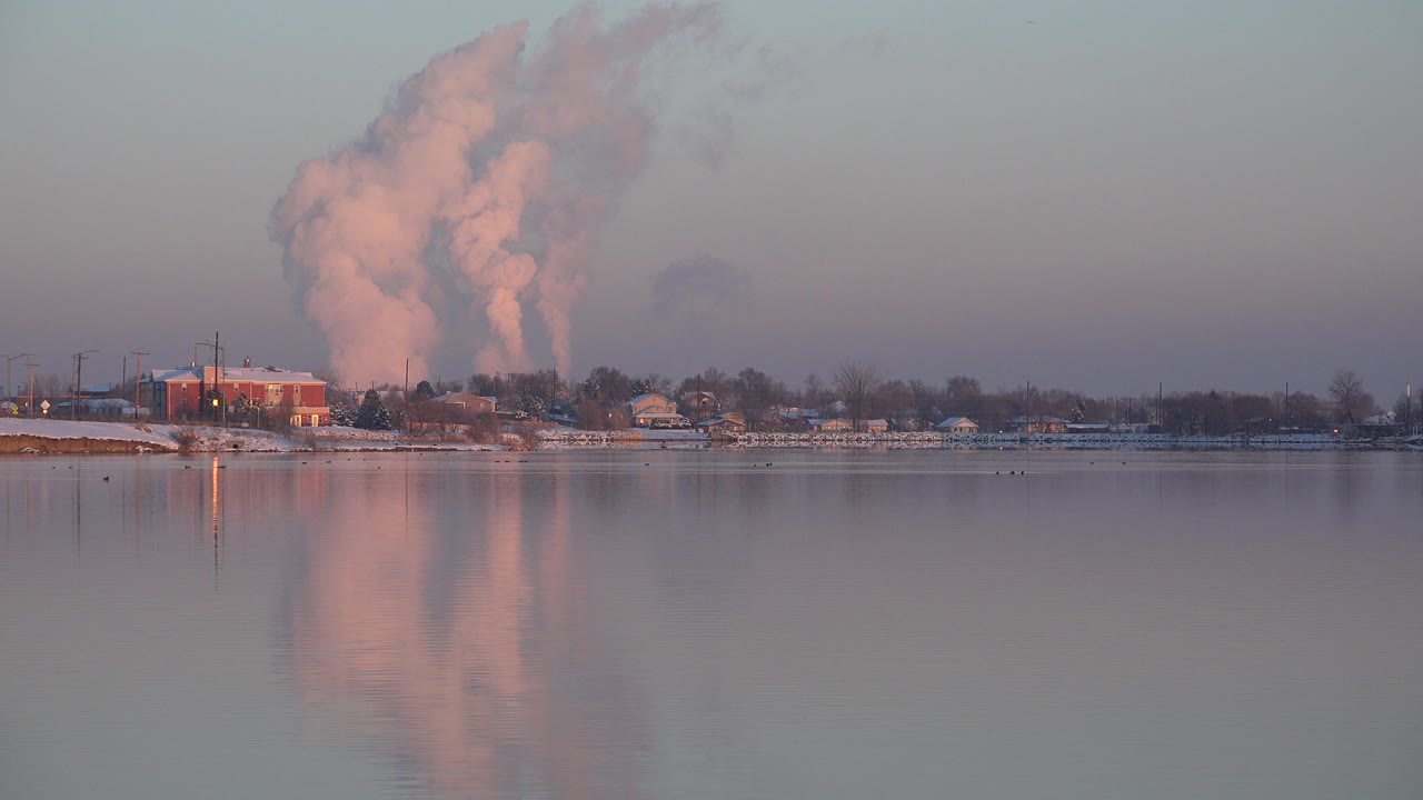 Wide view in Westminster, Colorado of a riverside industrial zone with a pink smoke plume rising behind trees, reflected in still water, with no visible people.