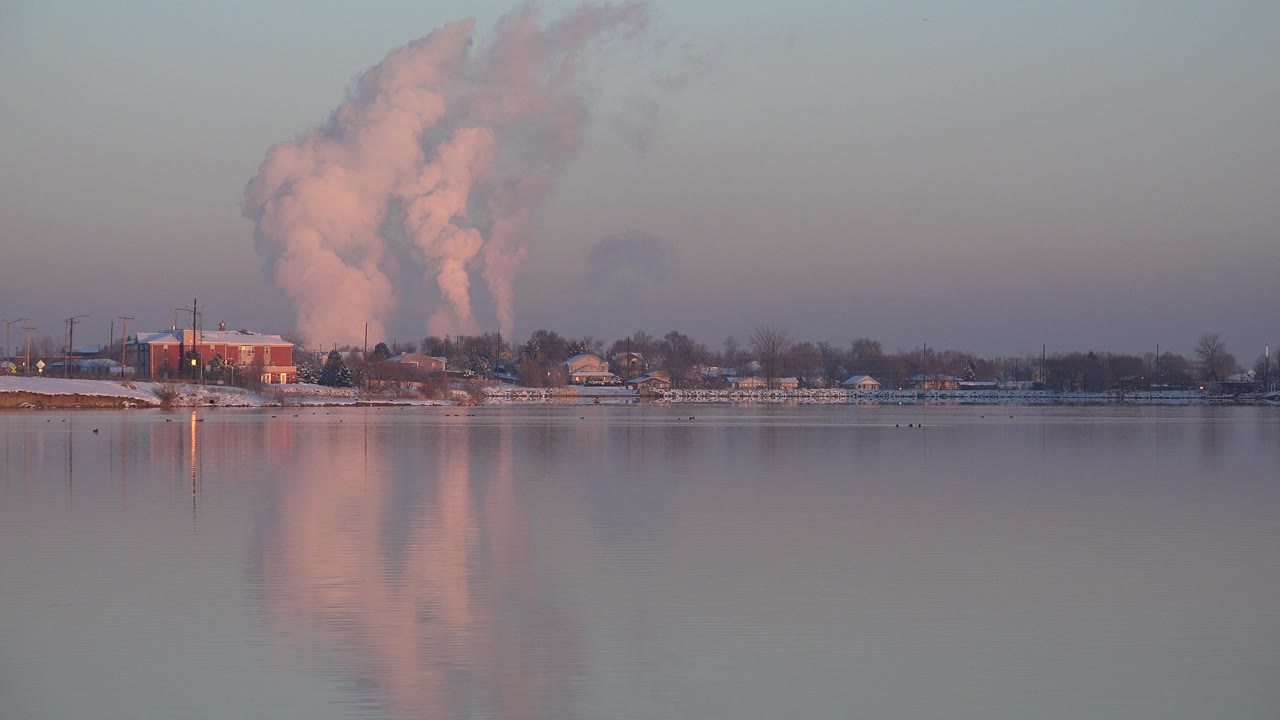 Westminster, Colorado riverside frame with factory structures and houses or small buildings along the far bank, a tall pink smoke plume, muted sky, and no visible people.