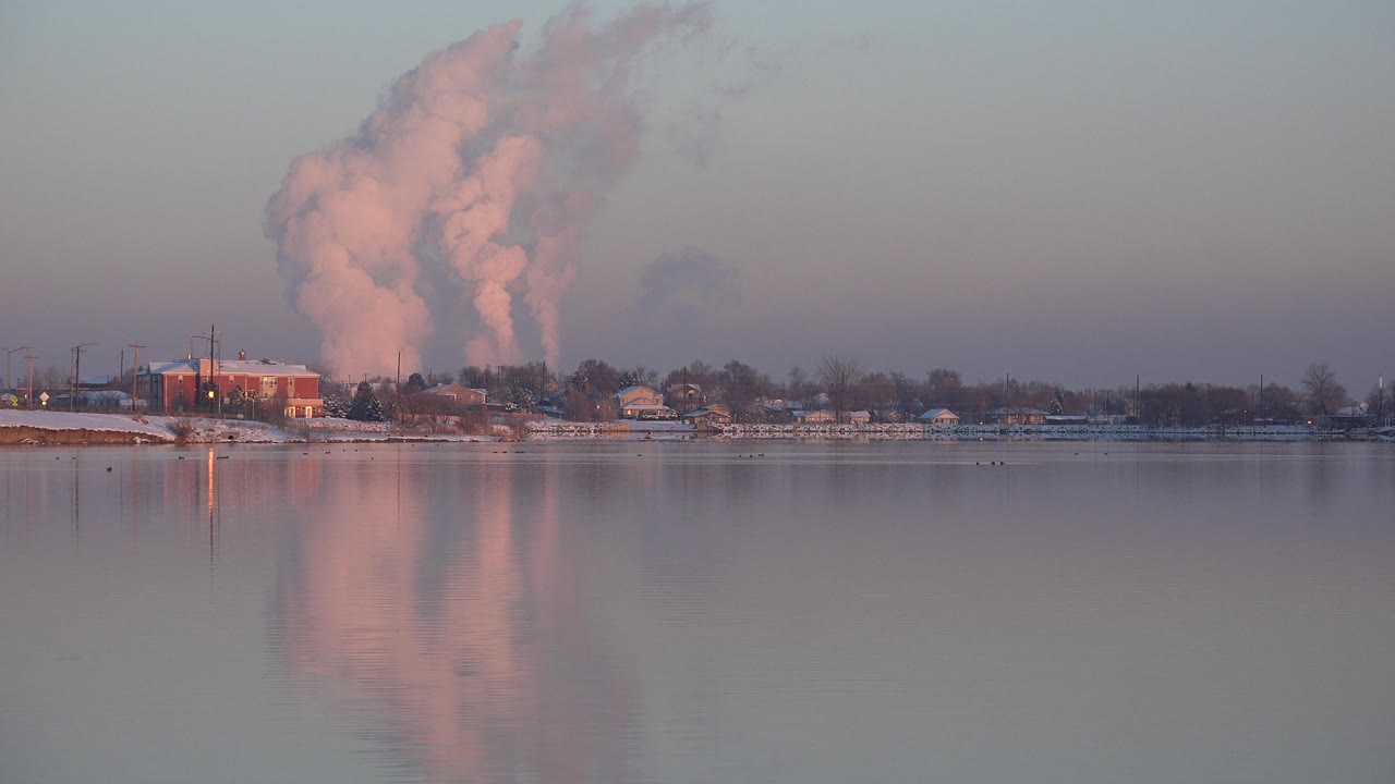 Factory area and pink smoke reflected in broad river water.