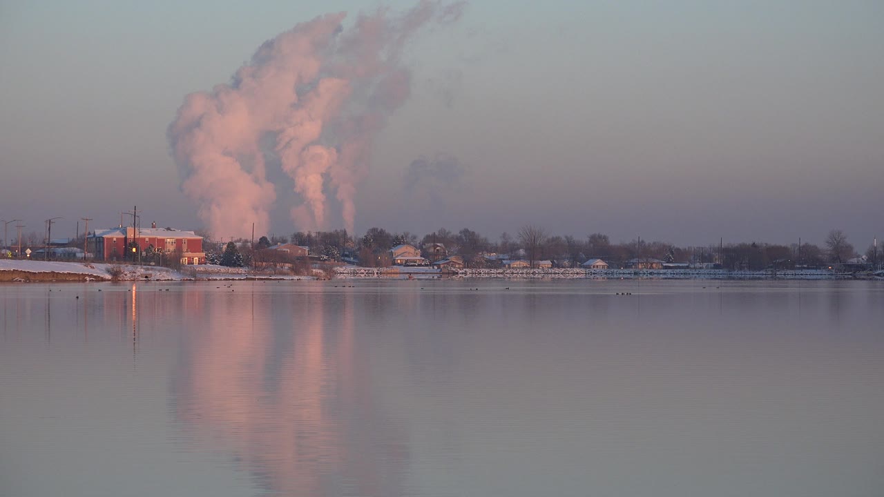 Wide Westminster, Colorado waterfront view at the start of the clip, showing snowy shore buildings, a large pink steam plume, reflected water, and no visible people.