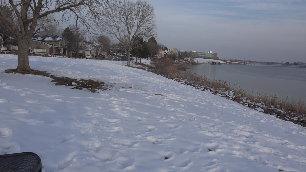 Wide winter view of a Westminster, Colorado lakeshore with snow-covered ground, bare trees, nearby houses, calm water, and no visible people at the start of the clip.