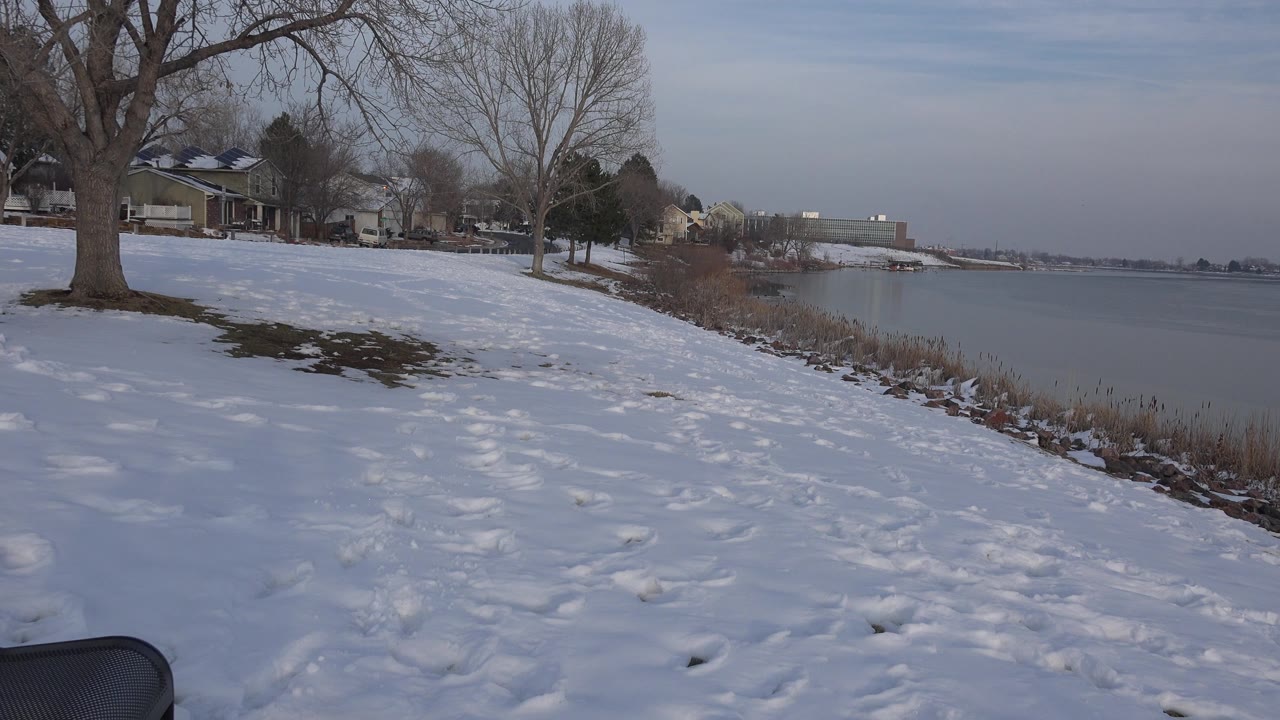 A low shoreline view in Westminster, Colorado shows snow, footprints, leafless trees, houses along the bank, muted water, and no visible people.