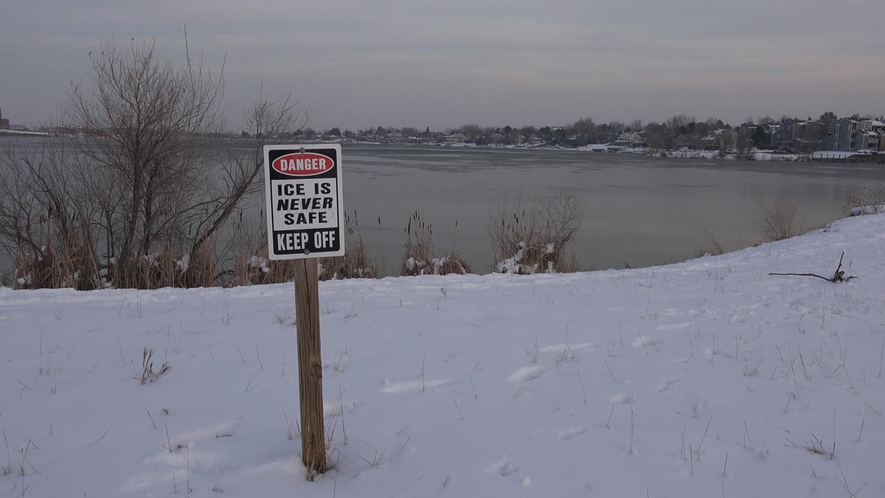 A red, black, and white danger sign stands on a wooden post beside a snowy frozen pond in Westminster, Colorado, with bare shrubs and no visible people.