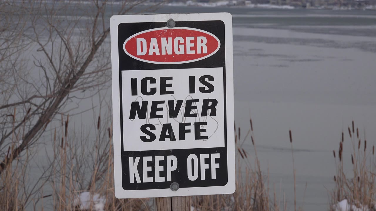 In Westminster, Colorado, a red and black Danger sign reading Ice Is Never Safe and Keep Off stands near a gray frozen lake with bare branches and no visible people.