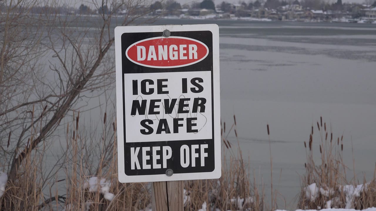 The warning sign remains large in the foreground with gray ice, shoreline reeds, leafless branches, distant Westminster homes, and no visible people.