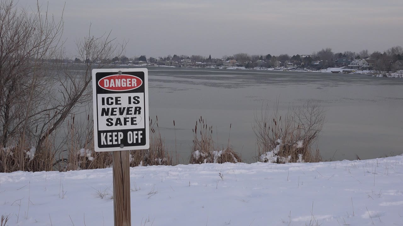 The unsafe ice warning sign remains prominent near the left side of the frame, with a flat gray lake surface, winter shoreline, and no visible people in Westminster, Colorado.