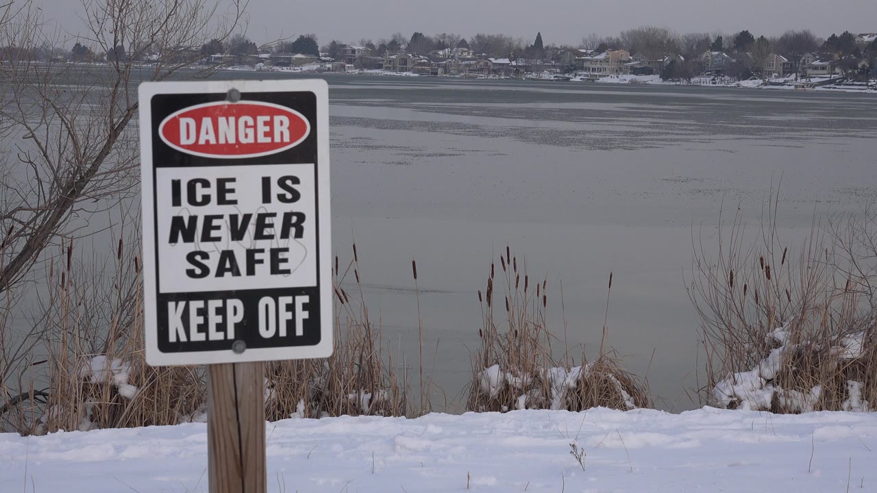 Danger ice warning sign beside snowy shoreline and gray water.