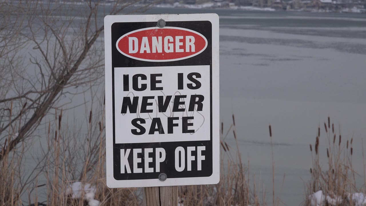 At 0:01, the sign remains large in the foreground while gray pond ice and winter vegetation fill the background, with no visible people.