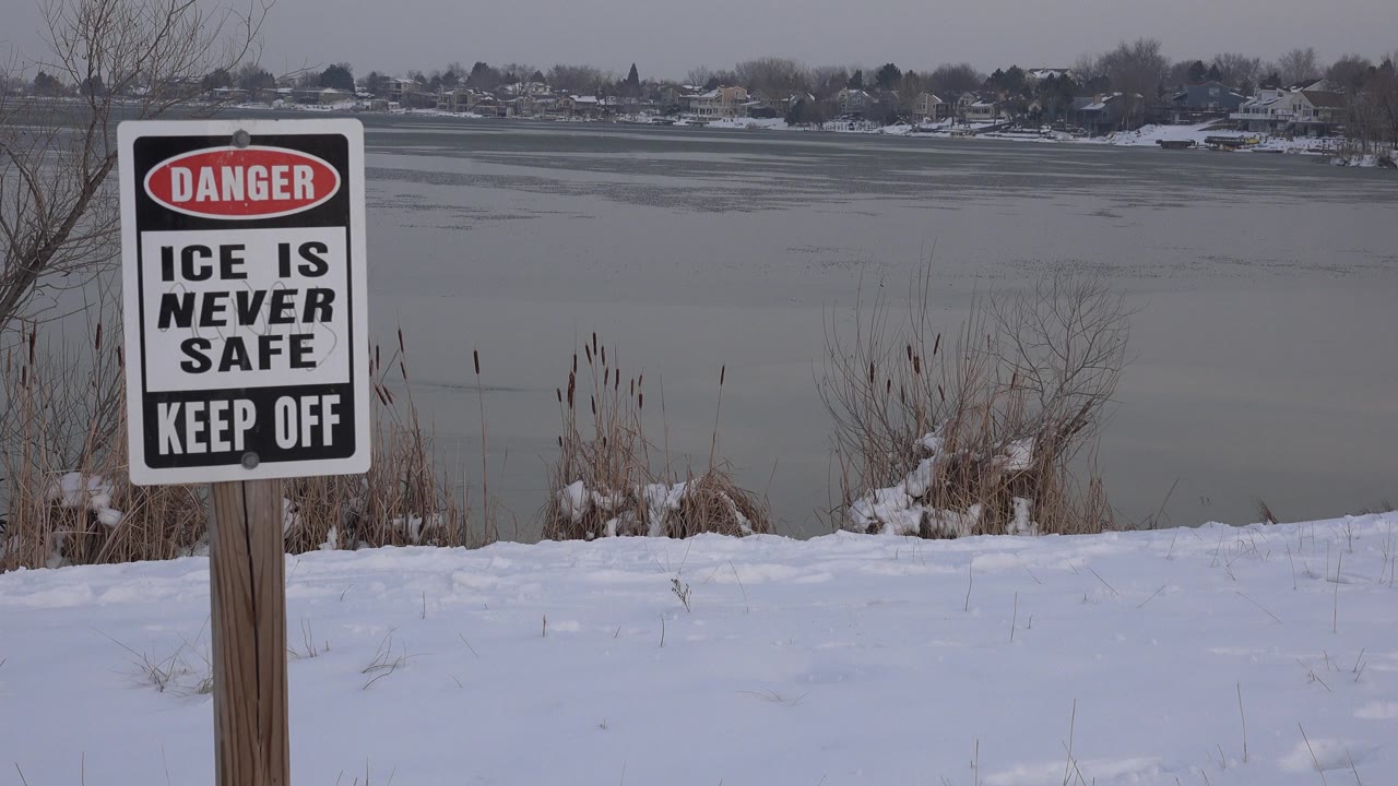 At 0:00, a danger sign reading ice is never safe and keep off stands at the left edge of a snowy Westminster, Colorado lakeshore, with gray ice-covered water and no visible people.