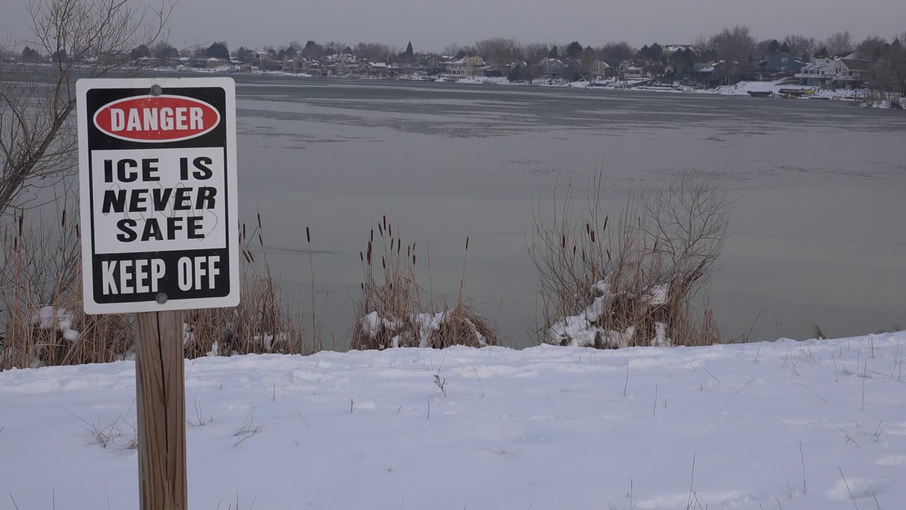 At 0:01, the ice safety sign remains near the left foreground on a snowbank, with reeds and the muted gray lake surface stretching toward distant Westminster shoreline buildings and no visible people.