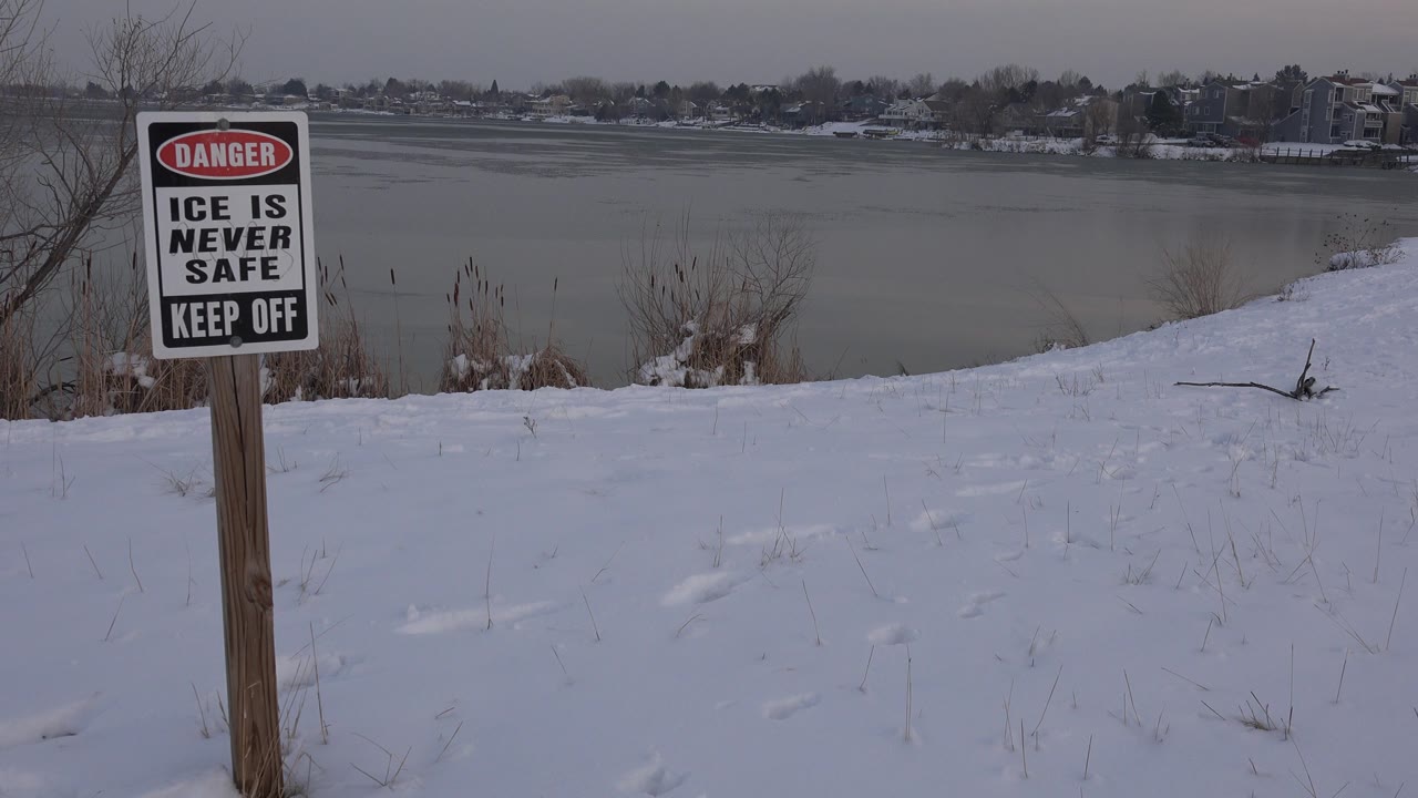 At 0:00 in Westminster, Colorado, a Danger Ice Is Never Safe sign stands in snowy foreground beside gray frozen water and a distant shoreline, with no visible people.
