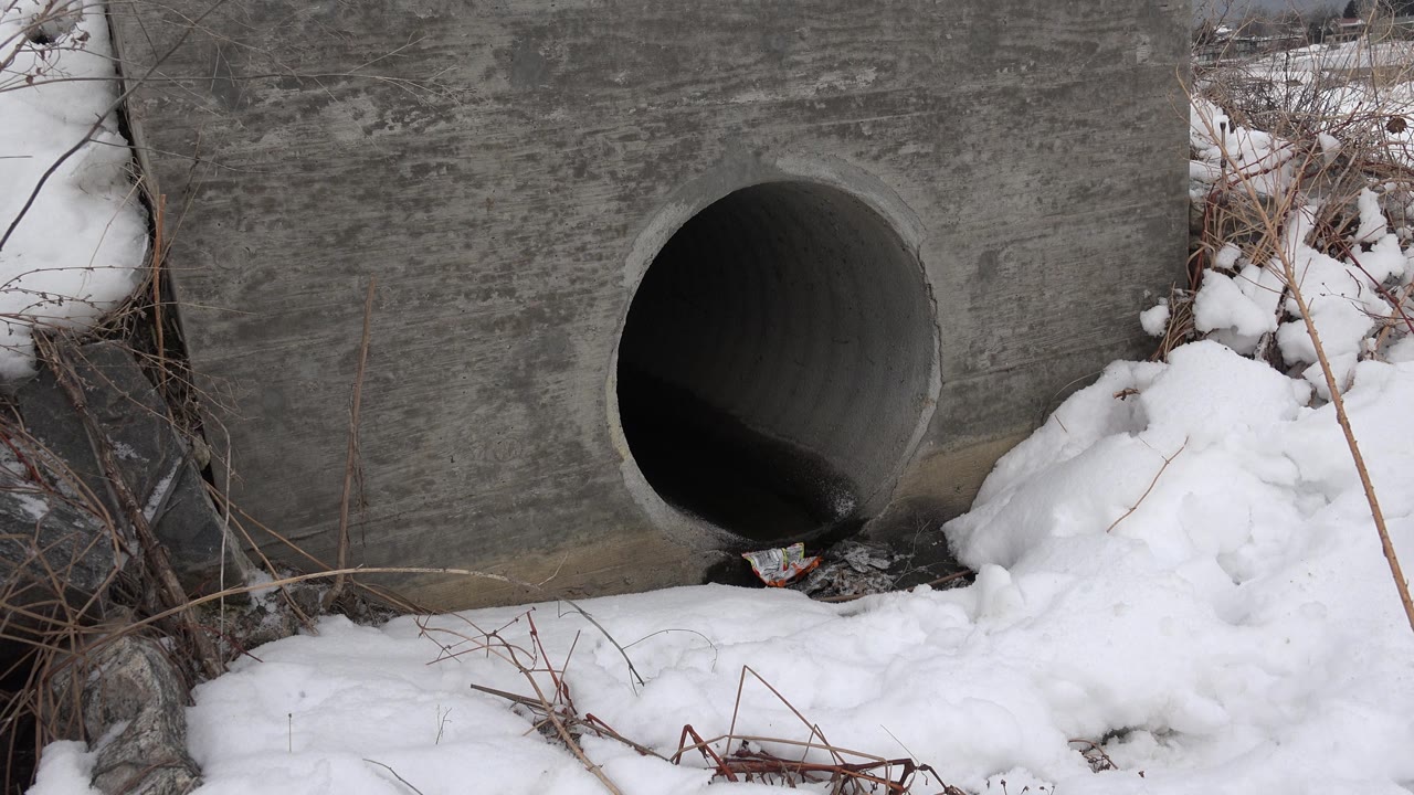 Gray drainage pipe set in concrete with snow and weeds in front.