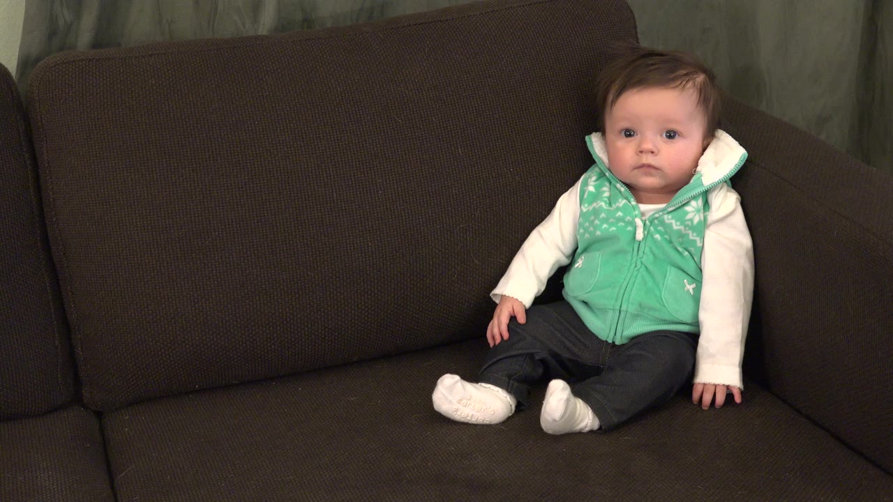Baby sitting upright on a brown couch in a mint green vest.