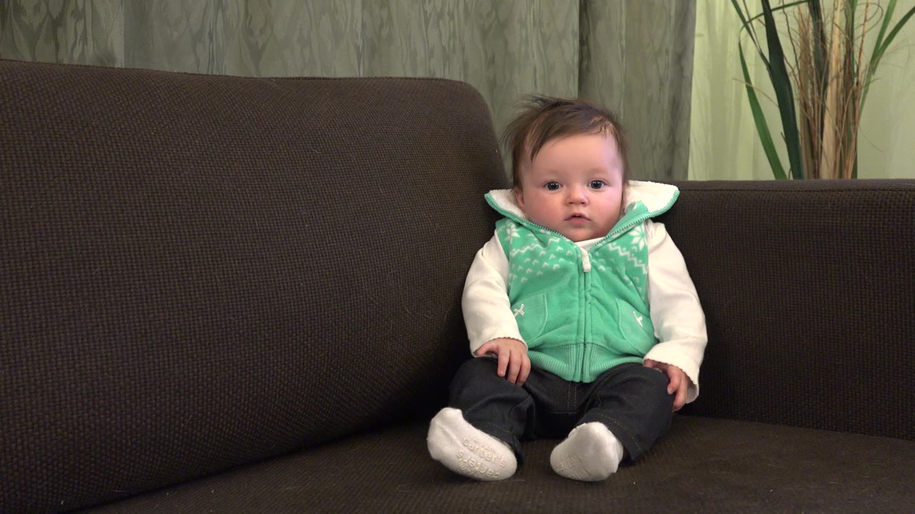 At 0:00, a baby sits upright on a dark brown couch in a Westminster, Colorado home interior, facing the camera with patterned curtains and plant stems behind.