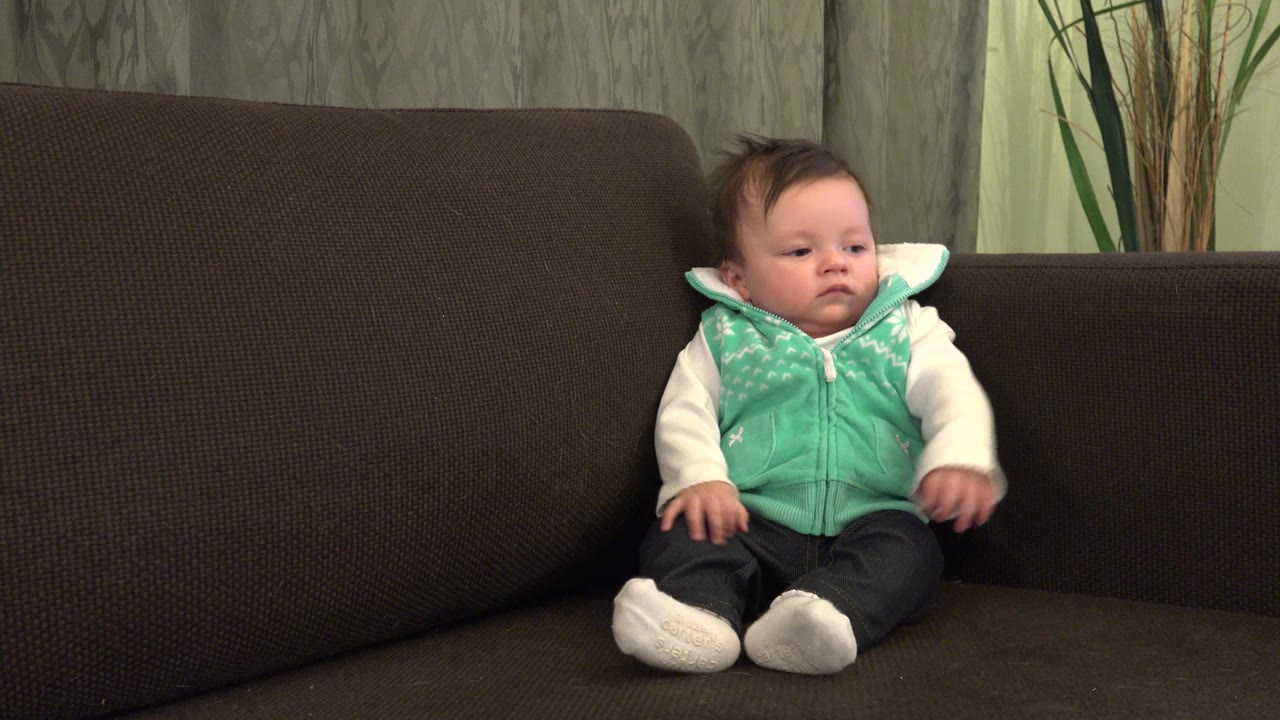The baby remains seated on the brown couch in Westminster, Colorado, with one hand raised slightly and the face turned toward the right side of frame.
