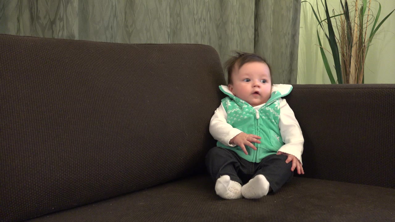 At 0:01, the baby remains seated on the dark sofa in Westminster, Colorado, with one hand slightly raised and the white shoes pointed toward camera.