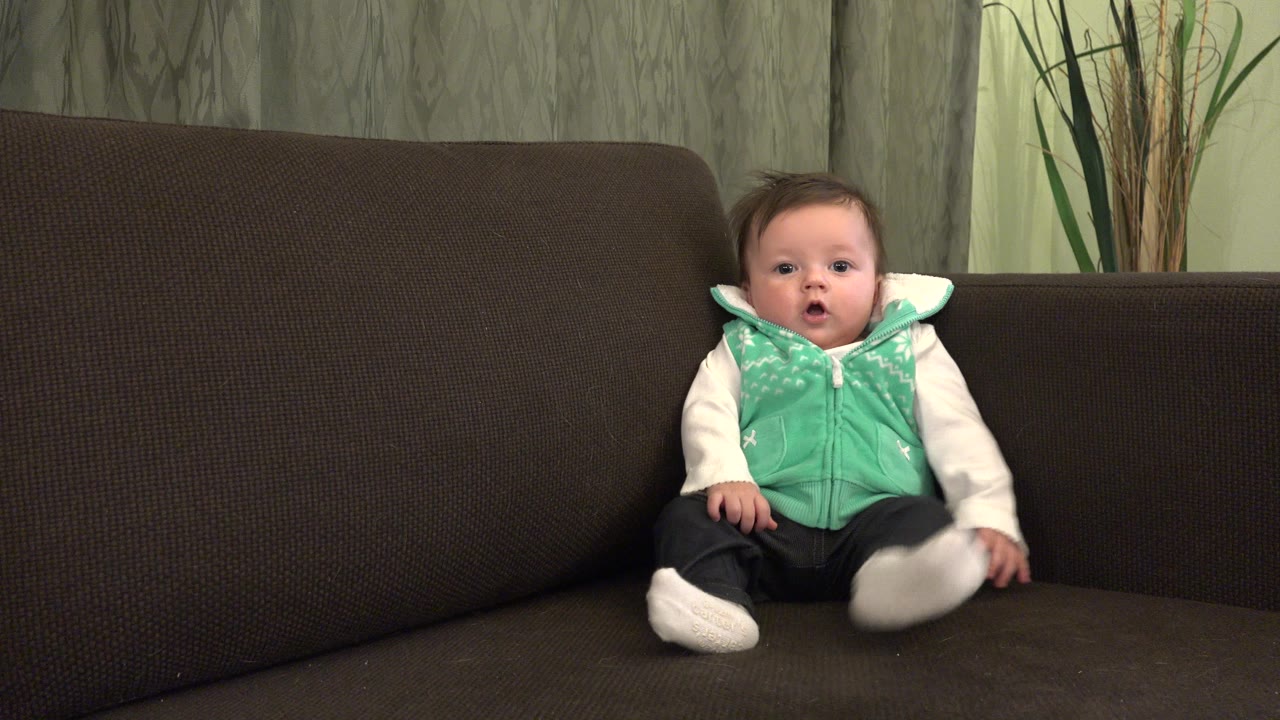 At 0:01, the baby remains seated on the brown couch in Westminster, Colorado, with one white shoe lifted forward and their face turned toward the camera.