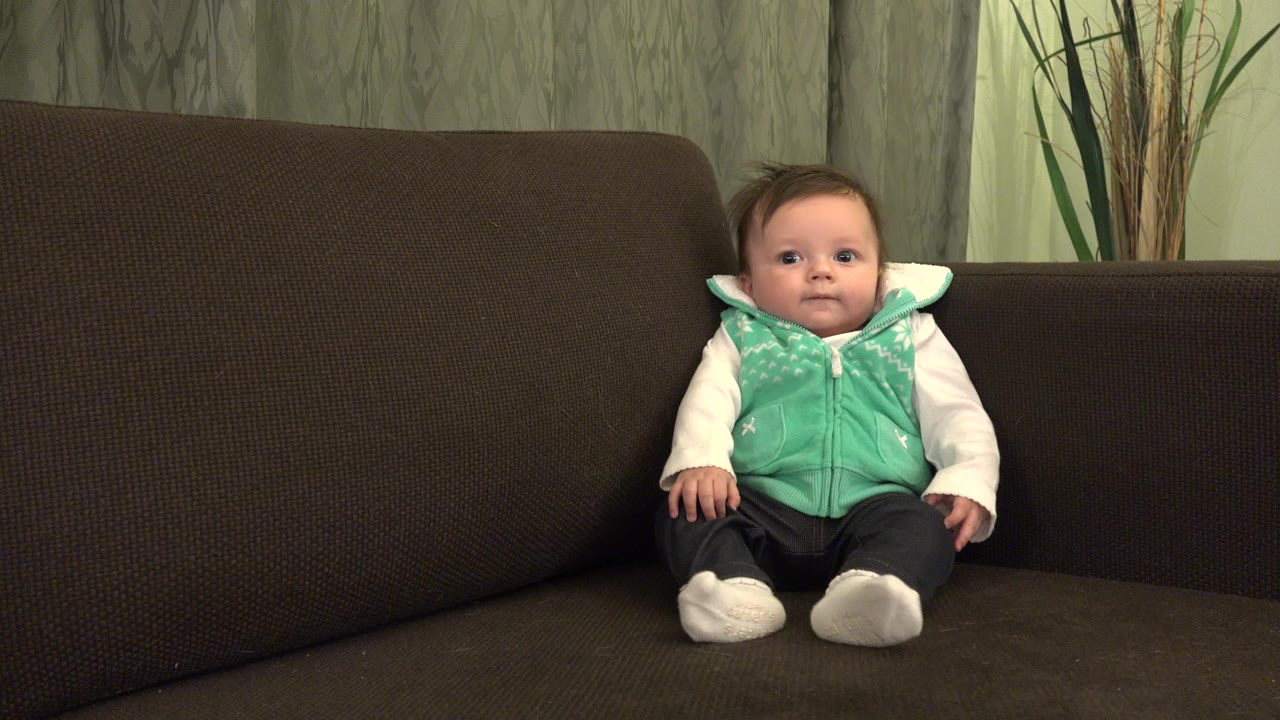 Baby sitting on a brown couch in a green vest.