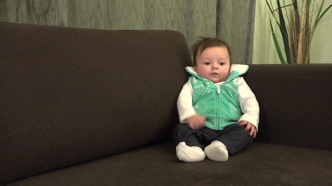 The baby remains seated on the brown couch in the Westminster, Colorado interior, with one hand lifted near the vest and the couch back filling most of the frame.