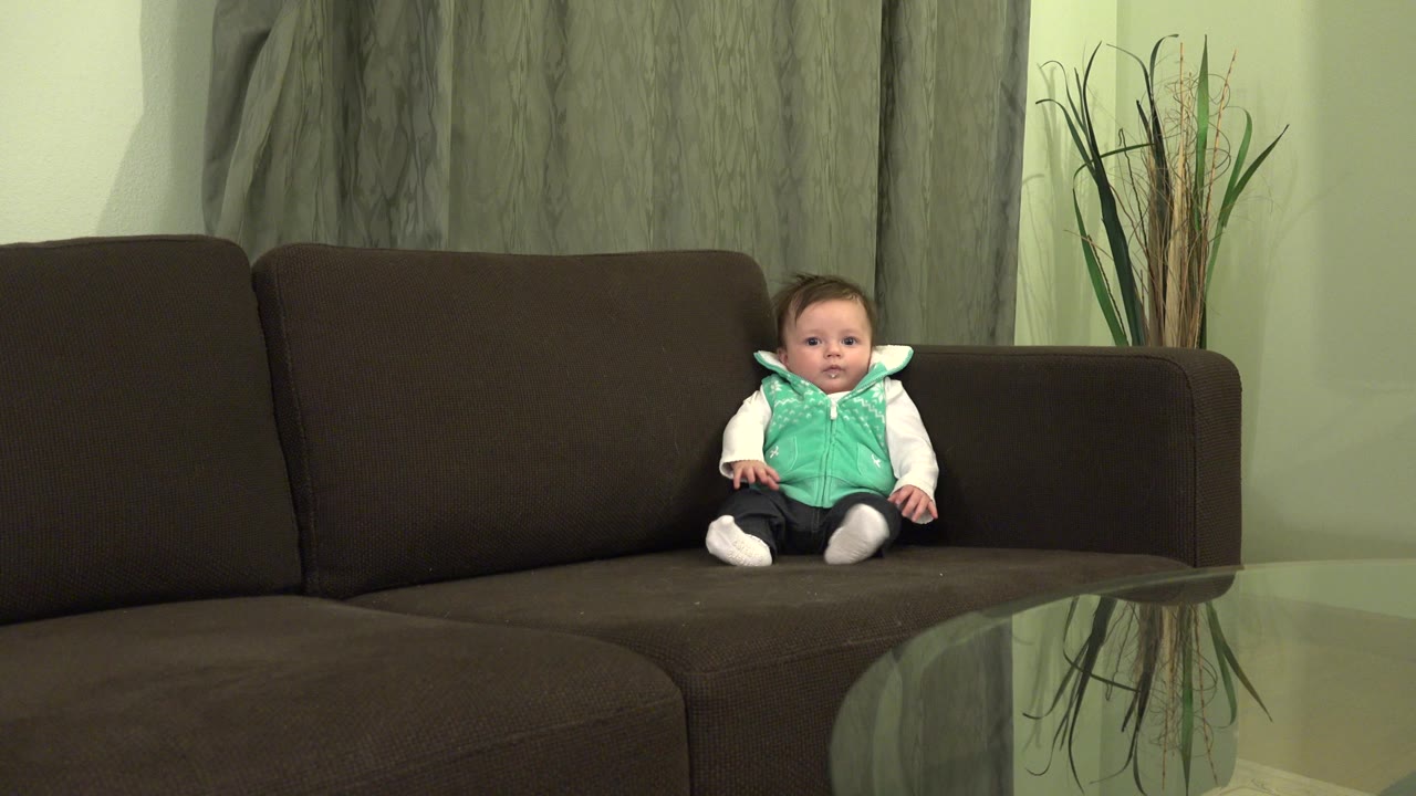 At 0:01, the infant remains seated on the right side of the couch, facing the camera with hands resting near the cushions and curtains behind.