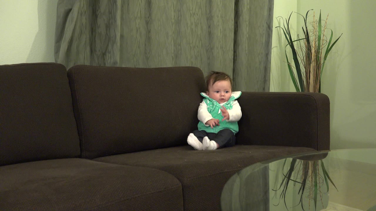 At 0:00, a baby boy sits near the right arm of a dark brown sofa in a Westminster, Colorado living room, with gray curtains and tall decorative reeds behind him.