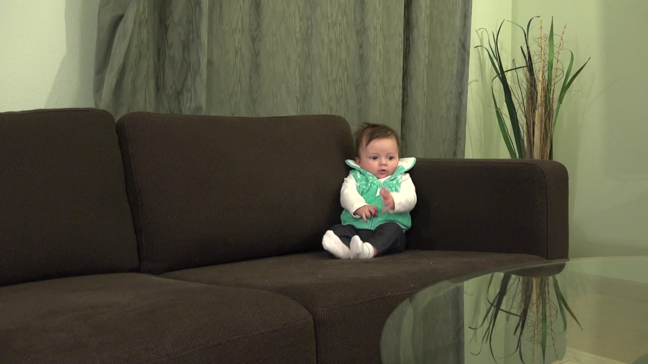 At 0:01, the baby boy remains seated on the dark sofa in the Westminster home, facing forward with the glass tabletop reflecting part of the room in the foreground.