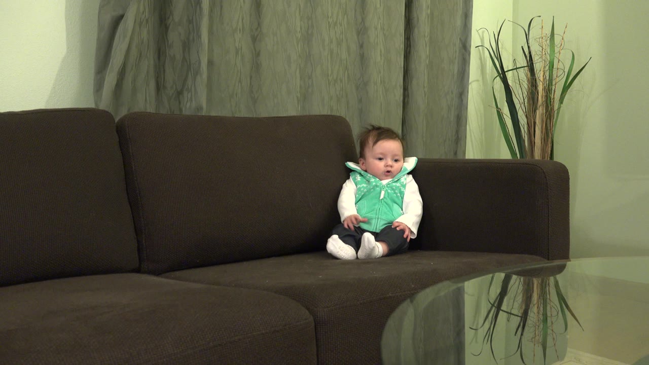 At 0:00, a baby sits upright on the right side of a brown sectional sofa in a Westminster, Colorado living room, with gray curtains and decorative reeds behind them.