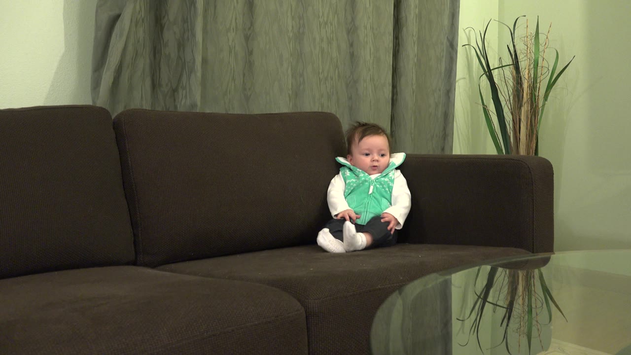 Baby seated near the right arm of a brown sofa.