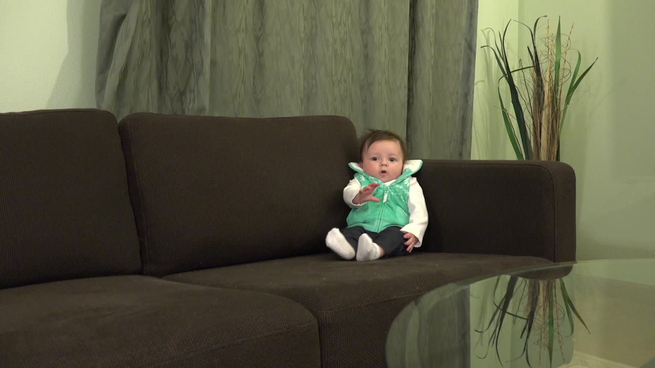 At 0:01, the baby boy remains seated on the couch in the Westminster, Colorado home, with one hand lifted near his chest and the glass table edge visible in front.