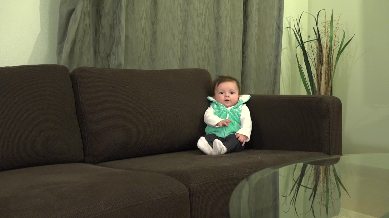 At 0:00, a baby sits upright on a dark brown sofa in a Westminster, Colorado home interior with green walls, gray curtains, and decorative reeds behind the couch.