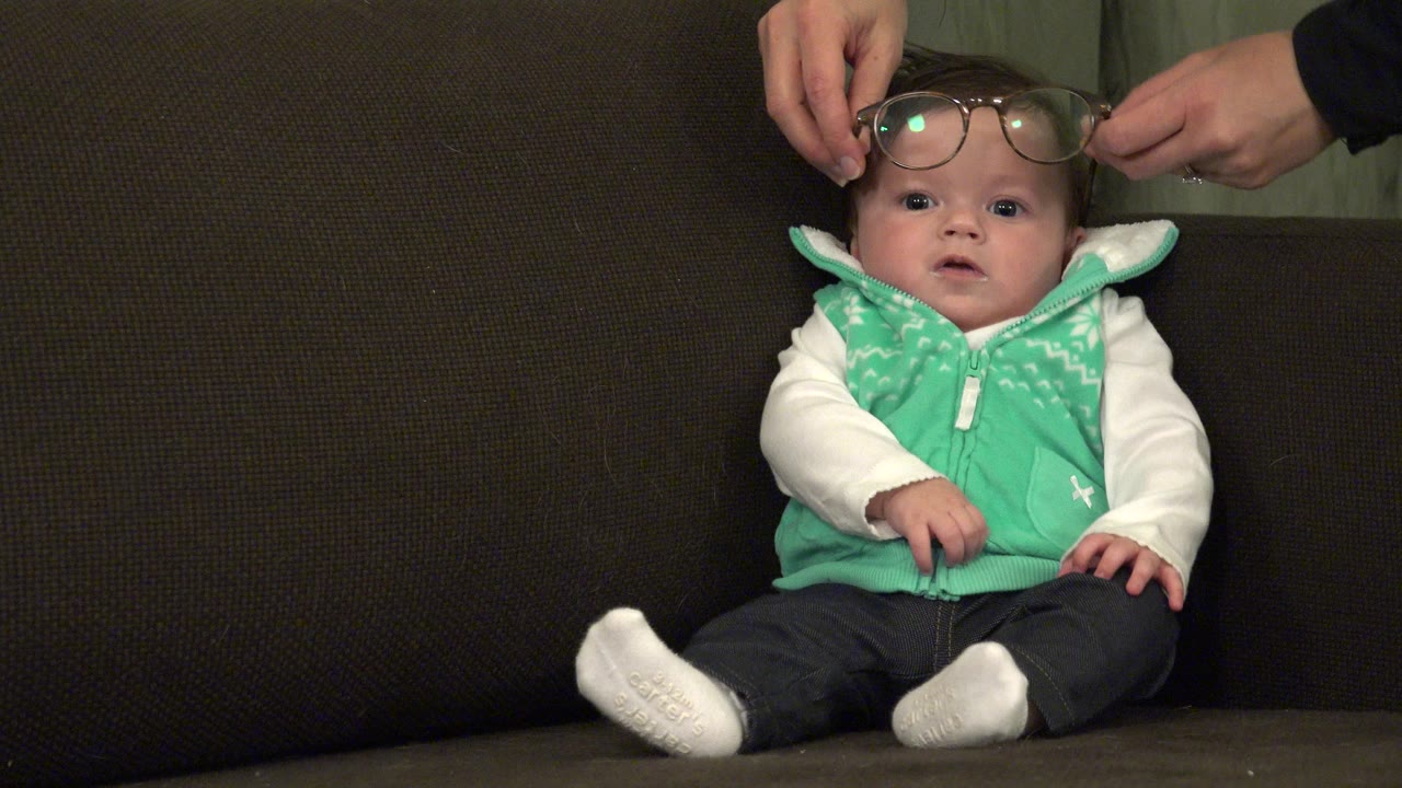 Baby seated on a dark couch as adult hands hold eyeglasses above the face.
