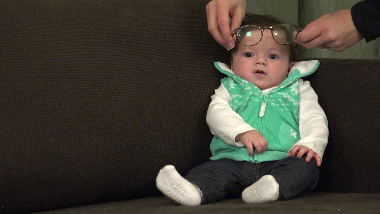 The baby looks toward the camera from the couch as adult hands continue positioning the eyeglasses across the forehead in Westminster, Colorado.