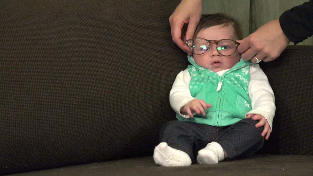 Infant sitting on a brown sofa as adult hands adjust large glasses.