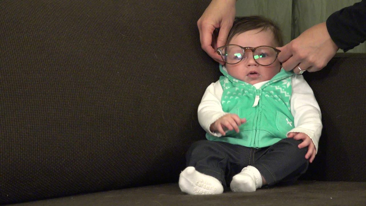 At 0:01 in Westminster, Colorado, the infant faces the camera on the brown sofa with oversized glasses reflecting indoor light as adult hands remain near the frames.