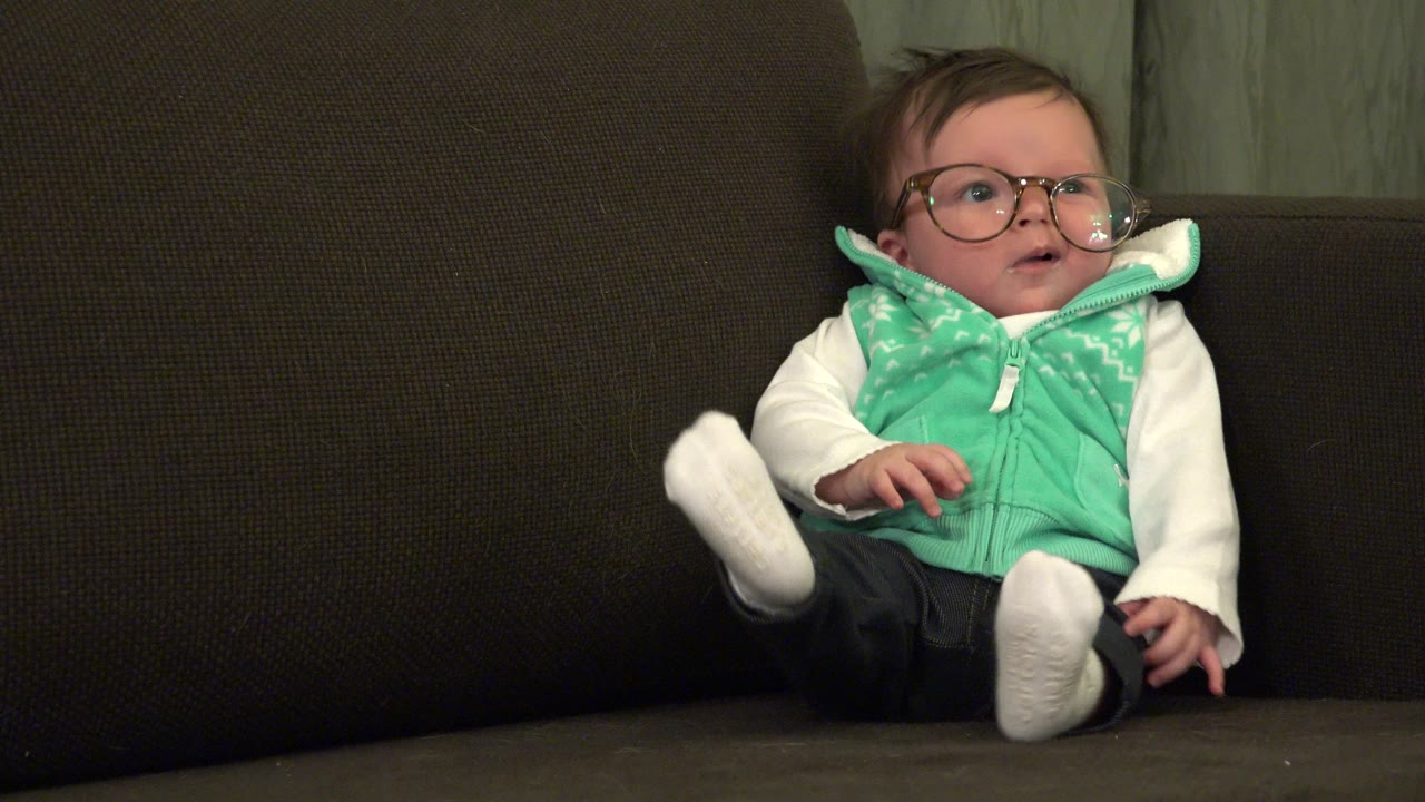 Baby wearing glasses and a green vest sits on a brown sofa.