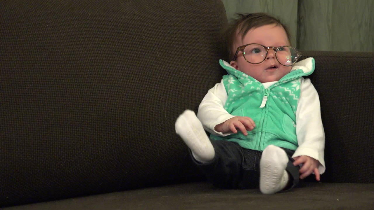 One second in, the baby remains seated on the brown sofa in Westminster, Colorado, looking slightly upward while wearing a mint green vest and large eyeglasses.