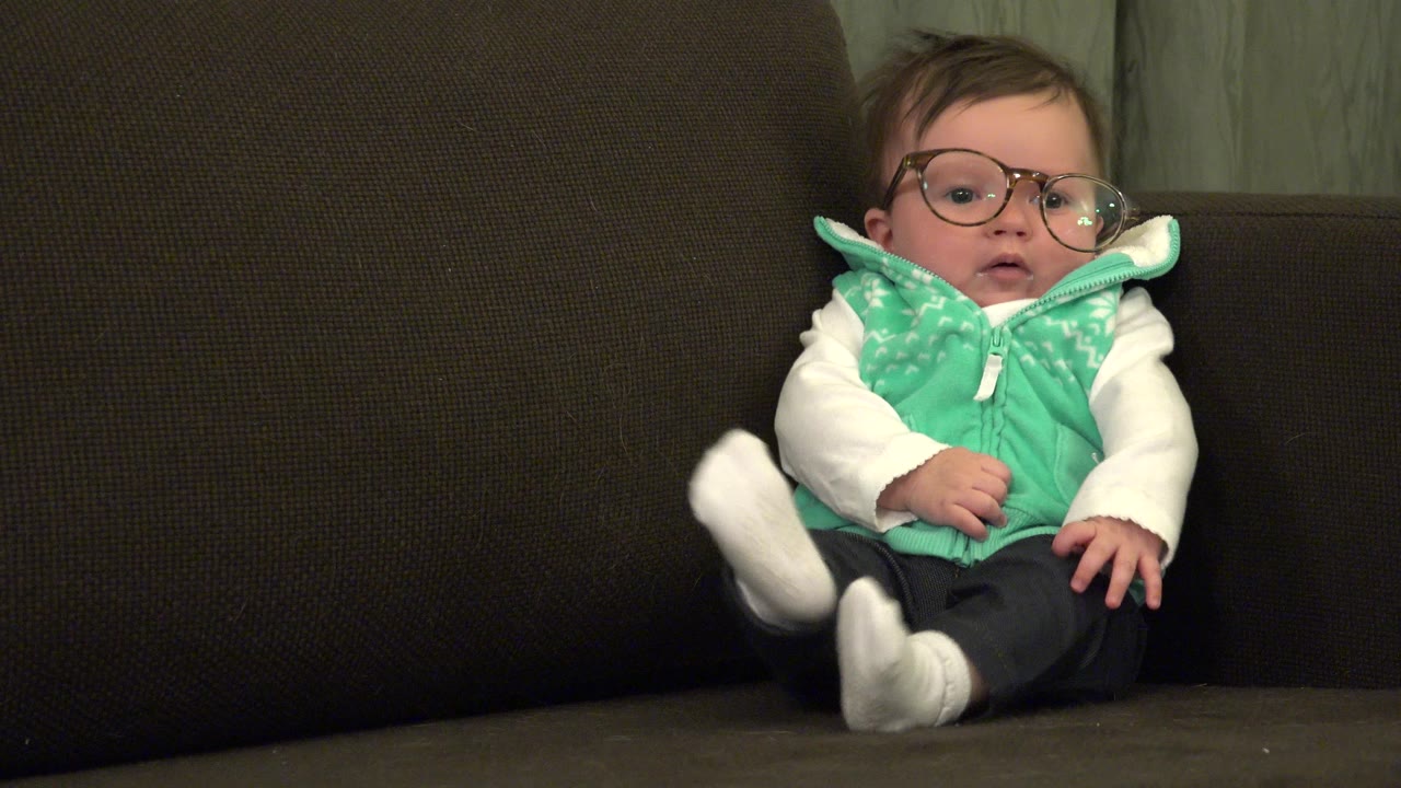 Baby in glasses sitting on a dark couch.