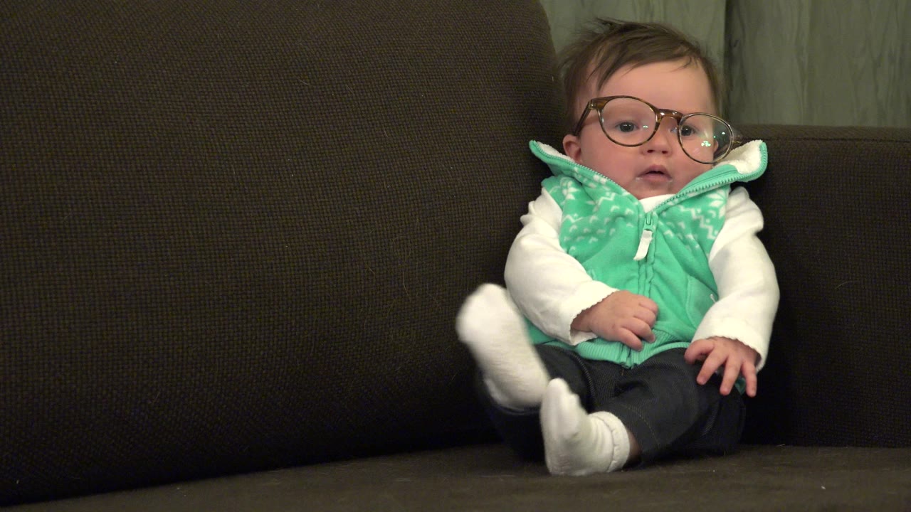 The baby remains centered on the couch in Westminster, Colorado, wearing glasses, a mint green vest, white sleeves, dark pants, and white socks.