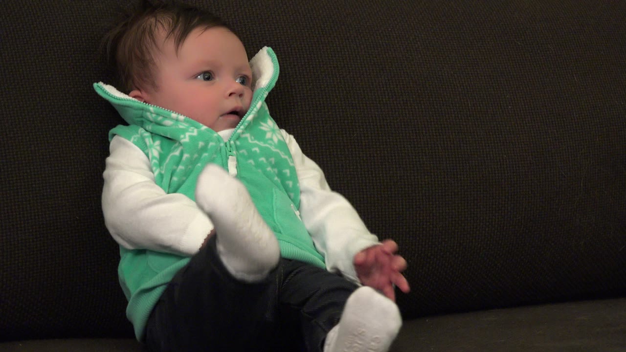 The Westminster, Colorado infant remains seated on the dark brown sofa, facing slightly right while both white-socked feet are lifted in front.