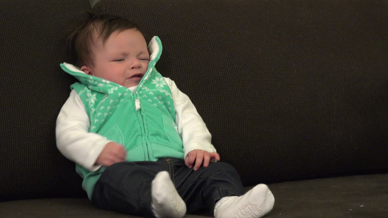 At 0:00 in Westminster, Colorado, a baby with dark hair sits on a dark sofa in a mint green vest, white sleeves, dark pants, and white shoes with eyes closed.