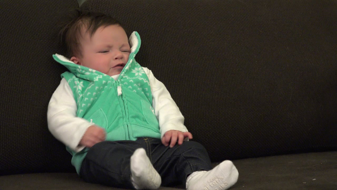 At 0:01 in Westminster, Colorado, the baby remains seated against the dark sofa back with eyes mostly closed and one hand resting near the lap.