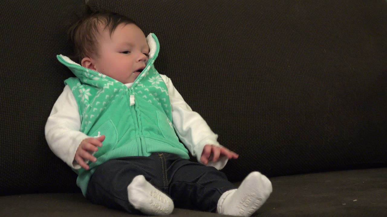 Baby on sofa turning head to the right with one hand lifted.
