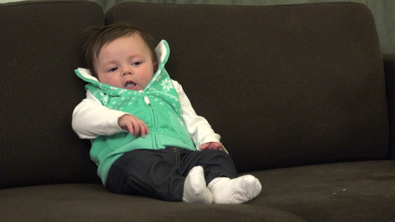 Baby sitting on a dark couch in a green vest.