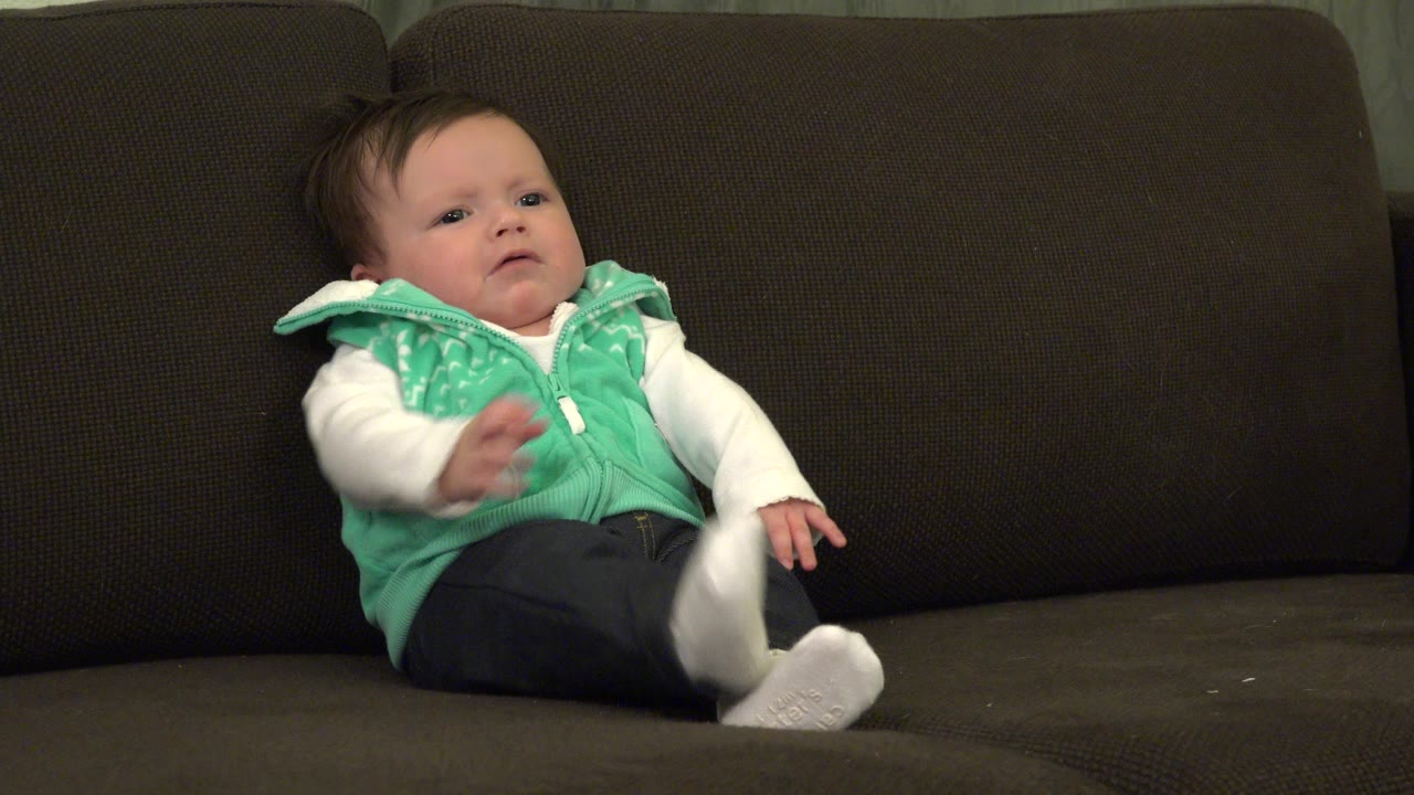 At the start of the Westminster, Colorado clip, a baby sits left of center on a dark brown sofa with one hand lifted and white socks facing the camera.