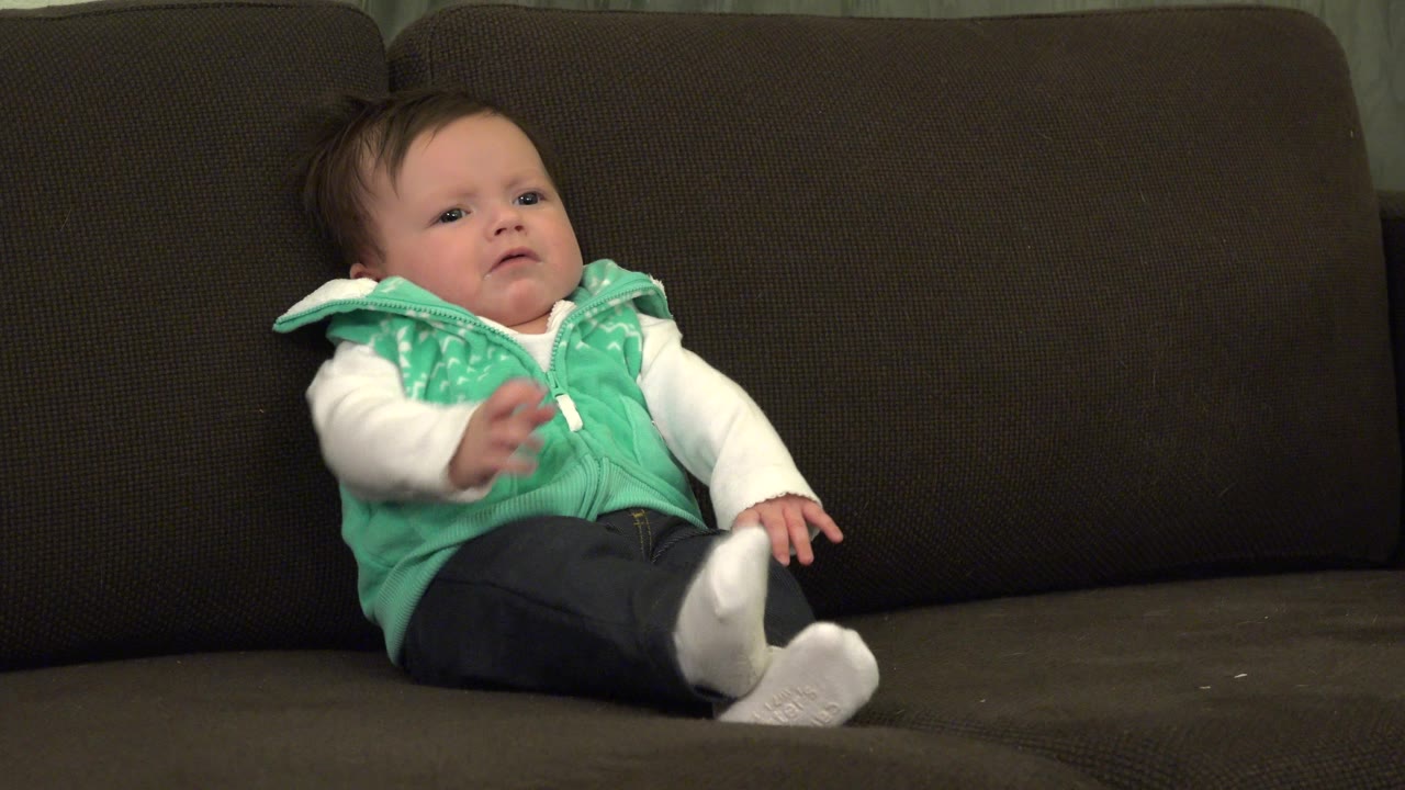 One second in, the baby remains seated on the dark sofa in Westminster, Colorado, with a small arm movement and a curious look toward the room.