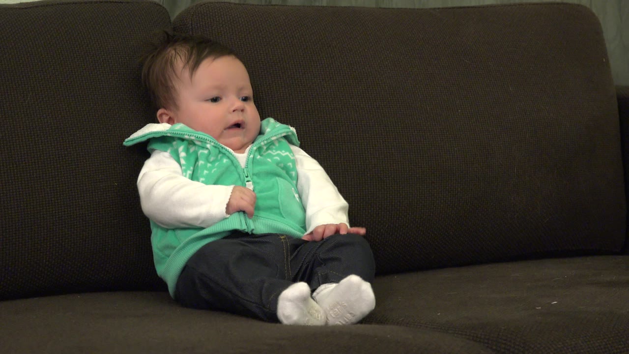 Infant in a green vest sitting on a brown sofa.