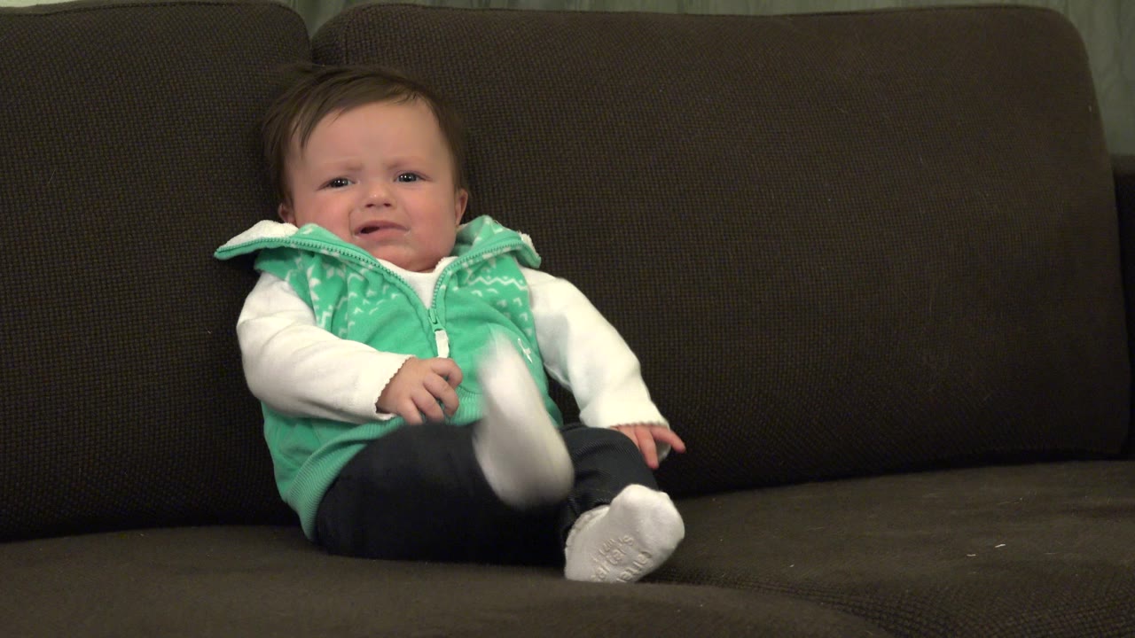 Baby sitting on a dark sofa with one leg lifted.
