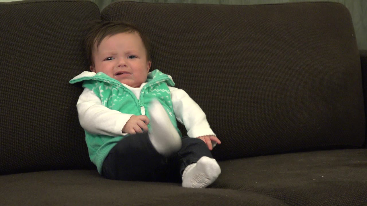 At 0:01 in Westminster, Colorado, the baby remains seated on the dark sofa while a white-shoed foot lifts forward and the arms stay near the body.