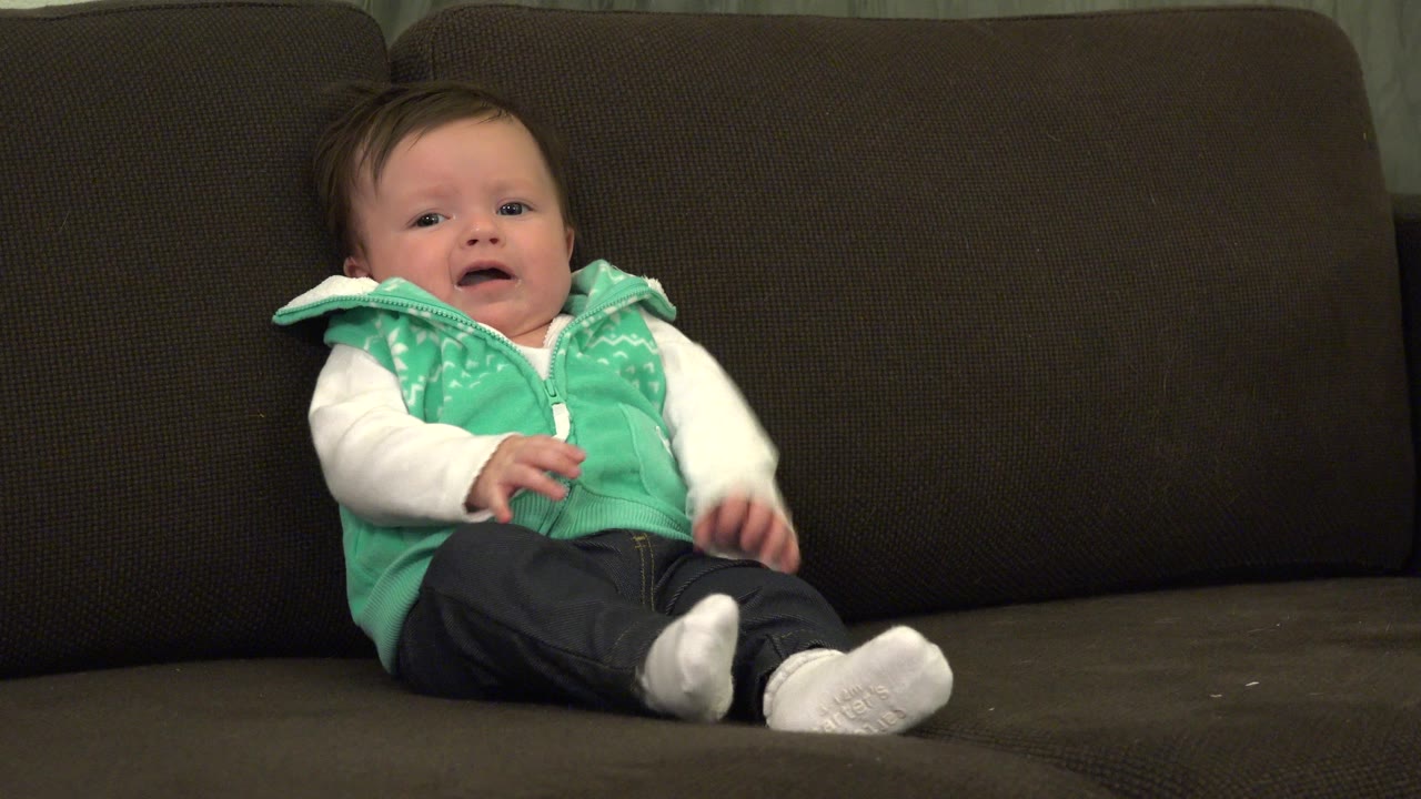 Baby sitting on a dark couch in a green vest.