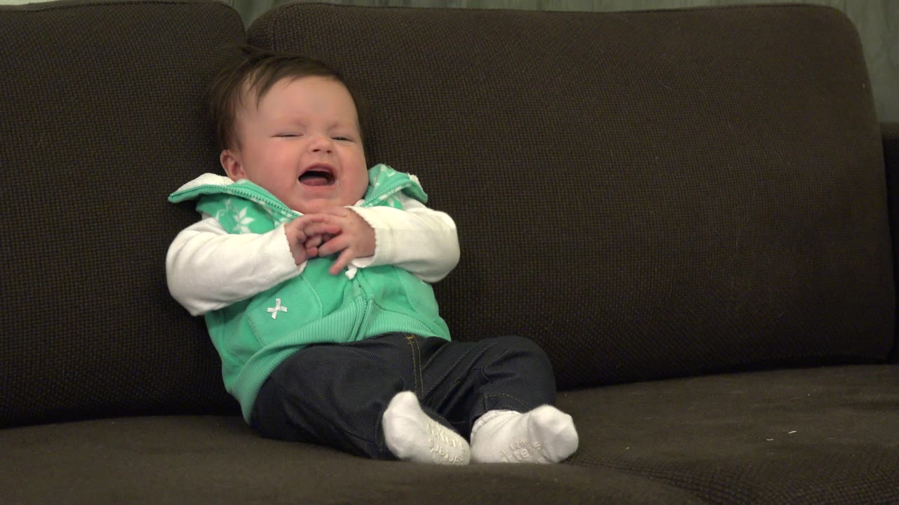 Baby crying on a dark sofa in a mint vest and white sleeves.